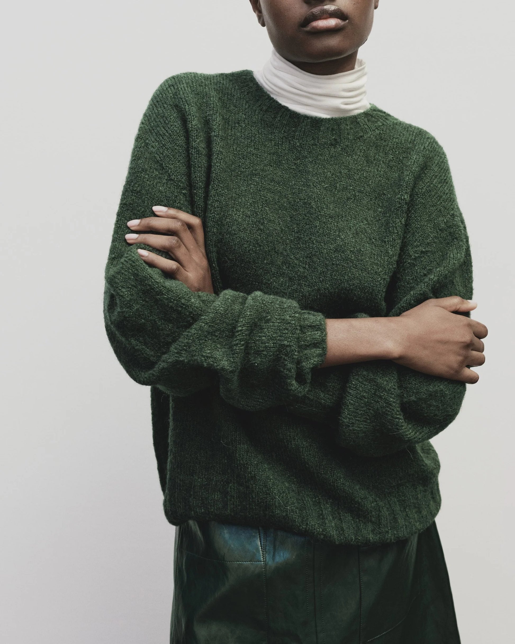 Person wearing a green sweater over a white turtleneck with arms crossed against a plain background